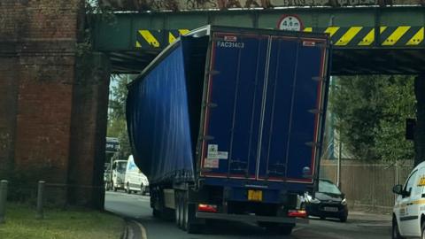 A lorry stuck under a railway bridge - there are yellow chevron markings at the top of the bridge - and the curtain on one side of the lorry is hanging off, with visible damage to the top of the vehicle.