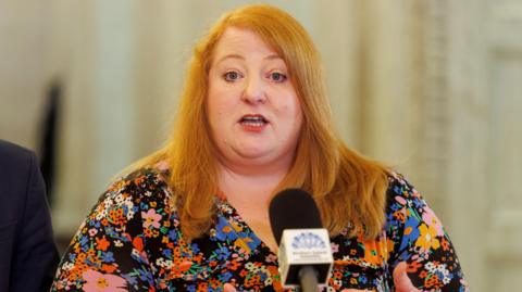 Naomi Long speaking into a microphone. She is wearing a patterned top and has long red hair.