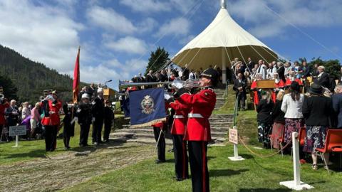 Trumpeters in red uniforms play in front of the hill, which is covered by a canopy, while dignitaries look on.
