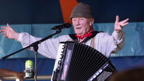 The Wurzels' Tommy Banner acknowledges the crowd. He wears a stripped shirt and brown hat with a red bandana round his neck. A cider sits in a drinks holder attached to his microphone.