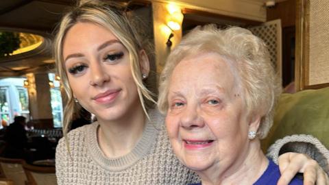 A picture of a female in her 20s with her arm around her grandmother. The younger female has blonde hair which it is tied up in a ponytail wearing a tan knitted jumper. She is wearing diamond studded earrings. The elderly lady on the right has blonde permed hair she is wearing a blue knitted jumper also she has a pair of diamond studded earrings.  