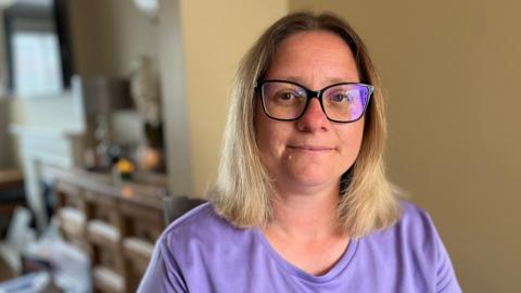 A woman with fair shoulder-length hair and wearing a mauve T-shirt and large, dark-framed glasses. She is sitting in a living room. Behind her on the left can be seen a blurred out chest of drawers. She is looking at the camera with a relaxed but serious expression. 