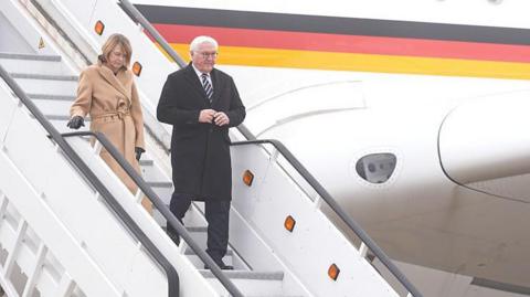 A man and a woman walking down the steps of a plane liveried in the German flag. She has fair hair and is wearing a long beige coat; he is in a long black coat, white shirt and black tie and has white hair.