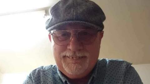 A man with a clipped grey beard and moustache smiles for the camera while wearing wire-rimmed glasses and a flat cap.