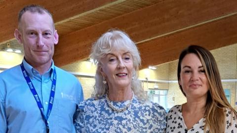 A man wearing a blue shirt and a lanyard, a woman with white hair wearing a blue floral dress and a woman wearing a white floral dress with brown hair stand with their arms around each other