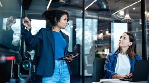 Stock image of a young woman holding an ipad standing next to a woman sat at a desk with a laptop. They are in an office or business setting and they are talking to each other.