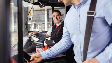 A man in a blue shirt taps a payment card on a contactless reader while boarding a bus. The bus driver , a man in a black vest, white shirt snd red tieis seated behind a transparent safety screen at the front of the vehicle, with the steering wheel and control panel visible. The image is taken inside the bus during daytime, with an urban street scene visible through the windows.