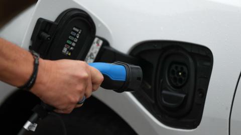 A picture of someone holding a charger. They are wearing a black wristband and holding a blue charger. It is a white vehicle with a black port.