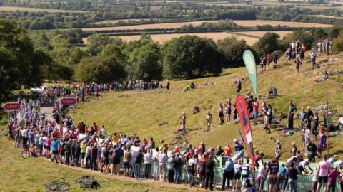 A road at the foot of a grassy hill is lined with spectators.