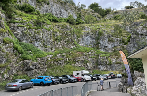 A view of a bend in the road in Cheddar Gorge - there is a row of parked cars opposite and a building to the right with the gorge rising in the background.