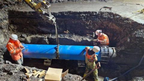 Three workers stand in a large hole in the ground, which has a large blue pipe running through it. The workers are wearing high-vis orange and yellow clothing and white helmets. The arm of a yellow construction vehicle can be seen at the top of the image, and it is holding the blue pipe in place.