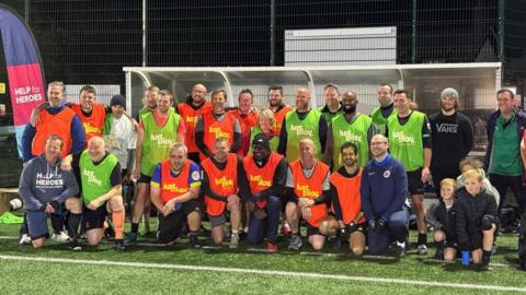 A group of football players wearing high visibility bibs in orange and green stand together as a team by the dugout on the side of the pitch. Former England player Emile Heskey is in the centre.
