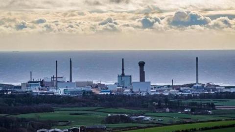 The cloudy sky, with the Sellafield nuclear site in the distance. It is made up of various large, tall buildings. Rolling fields can be seen in front.