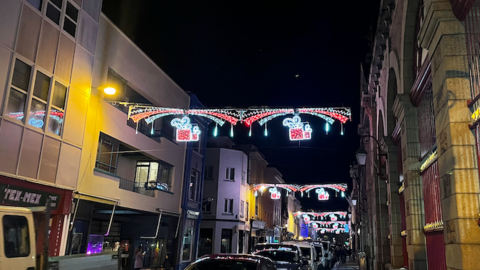 Red and white Christmas lights depict falling snow and wrapped presents hang above a road in St Helier which is next to the market.