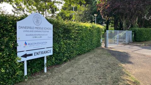 A view of a school building. The sign in front says Danesfield Middle School. There is a green hedge and a school gate.
