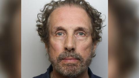 A police mugshot of Chris Brain. He has curly, swept back brown hair and a grey and brown-flecked beard.