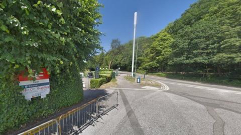 Ascot railway sign on left set in green hedge ascending road and footpath lined with leafy green trees 