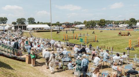 A landscape photo of spectators, several with straw hats on, sat around metal chairs and tables by a field with equestrian equipment and a horse and rider in mid-movement in it.