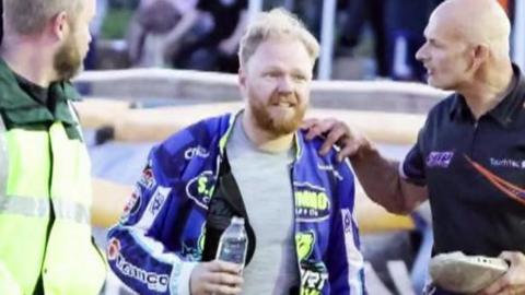 Rider Cameron Heeps walking away after the crash. His team are surrounding him. He is holding a bottle of water.