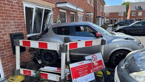 Red and white barriers fence off the area outside the grey car in front of the smashed glass panels of the ground-floor window