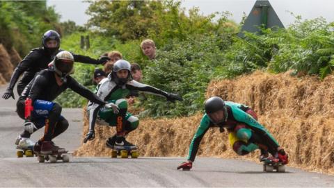 Five skateboarders coming down a road in helmets with hay bales lining the road.