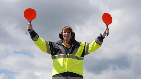 Angela Rayner outside wearing a yellow high viz jacket and holding what looks like a red badminton racket in each of her raised arms as she helps a plane manoeuvre at Stansted airport. She is smiling wearing eye defenders and has a cloudy blue sky behind her. 