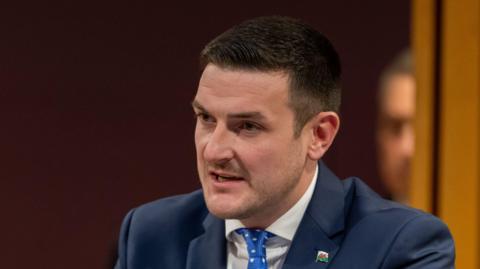 ames Evans is wearing a blue suit, blue tie and white shirt, in front of a dark background. He is looking away from the camera.