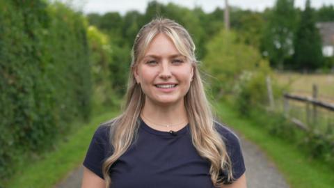 Wearing a navy blue round-necked T-shirt, Amy Thompson is standing on a countryside path surrounded by hedges and trees. Amy, with long, blonde hair, is smiling directly at the camera.