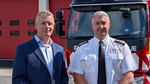Luke Pollard and Gavin Ellis stood in front of a fire engine parked in front of the fire station. Pollard is wearing a blue shirt and blue suit jacket. Ellis is wearing a white shirt with a black tie. They are both smiling at the camera.