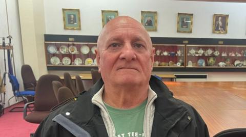 A bald man wearing a green T-shirt with a grey hoodie and then a black coat over it, stares into the camera. Behind him is a wooden table with glass display cases of pottery on the wall.