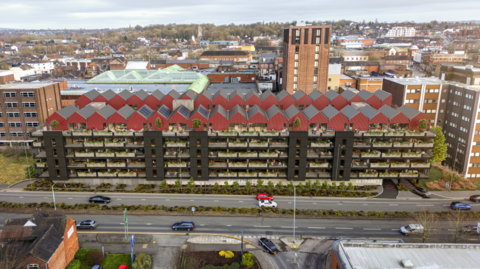 Artist impression of the apartments in place of the car park. Small, red roofs sit atop the former car parks with apartments on many levels below with green indoors or outdoor plants visible from the front of the building. The building is next to a road in the town centre with taller properties surrounding it.
