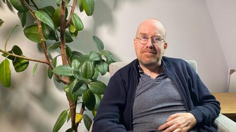 A man sat on an armchair next to a large rubber plant