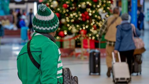 A traveler in an airport wearing green is walking away. Other travelers are in a blurred background along with a Christmas tree with gold and red ornaments and white lights.