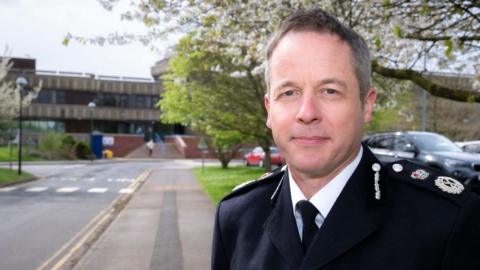 A head and shoulders view of Paul Gibson, the chief constable of Lincolnshire Police, wearing a dark uniform jacket with rank insignia on his shoulders, a white shirt and black tie. In the background is a large police station with dark grey concrete walls and dark-glass windows.