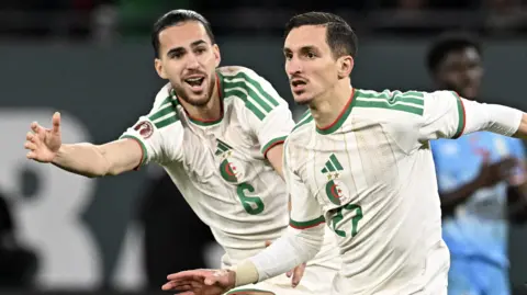 Adil Boulbina, wearing a white Algeria shirt, celebrates a goal alongside team-mate Ramiz Zerrouki