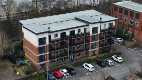 An aerial view of a multi-storey housing complex, featuring a flat roof, four storeys, and a number of cars parked in a car park running along the front of a building. Trees can be seen behind the building. 