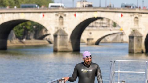 A swimmer emerging from the Seine in Paris wearing a wetsuit and a lavender swimming cap, with goggles over the top of it