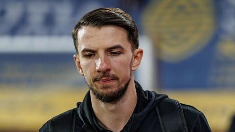 Dan Barlaser walks along a touchline in a black tracksuit with the strap of a bag over his left shoulder