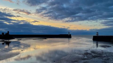 Mainly cloudy sky over the coast with a pier and a lighthouse in the distance 