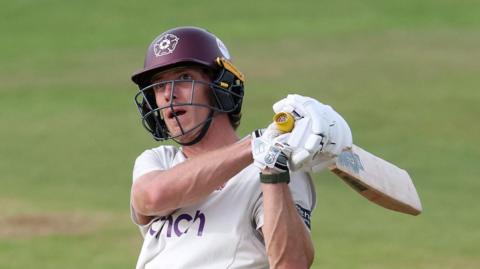Calvin Harrison of Northamptonshire swings a cricket bat during a County Championship match. He is wearing a white Northamptonshire jersey and a claret helmet with the Northamptonshire rose emblem.