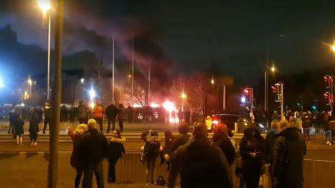 Crowds of people around on a street looking towards a fire in the distance