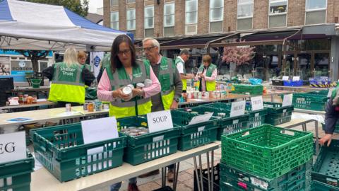 People dressed in high vis jackets with the words Shelter Trust on them are standing amongst trestle tables with green baskets containing tins. Each one is labelled with a different category, including soup and meat. A woman in the foreground is holding several tins of donated food.