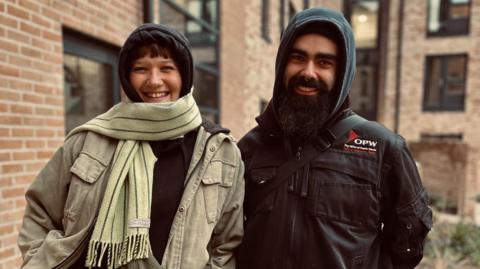 A young couple in their thirties stand outside a block of new apartments. They are both smiling. Chris has a dark beard and is wearing a dark jacket with a hood.  