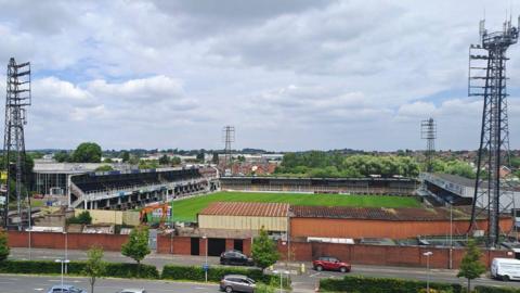 A football ground with a grey sky overhead. There are four large pylons with floodlights on them, one at each corner of the ground.