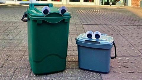 Two bins on concrete. One is large and green and the other one is smaller and blue. They both have googly eyes stuck on them 