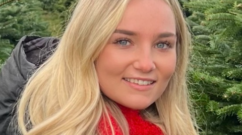 Emily is standing in a grove of Christmas trees. she is wearing a grey parka and has a red scarf tighed at her neck. She has long wavy blonde hair, blue eyes and is wearing light makeup. 