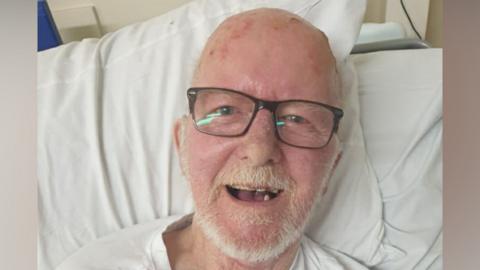A man with white hair and glasses in a hospital gown lying in a hospital bed. 