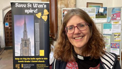 Rev Ruth Clay smiling at the camera. She has long brown hair over her shoulders, parted in the middle. She has purple rimmed spectacles and is wearing a blue and white hoodie over a floral top. Underneath her white 'dog' collar and black shirt can be seen. Behind her is a 2m (6ft) high display sign about a campaign to save the spire.