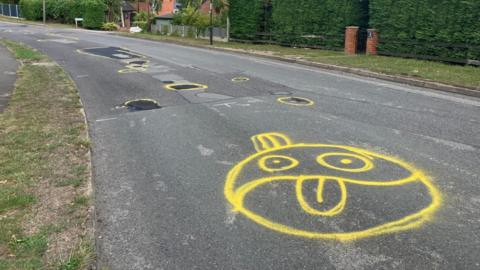 Circles in yellow spray paint around potholes which have been filled in along with a cartoon face with it's tongue out.