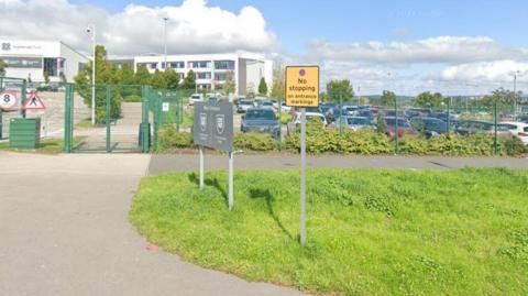 The outside of Co-op Academy Grange in Bradford. Picture is a modern-looking school behind a green metal gate and fence, while a 'no stopping' roadsign stands in the foreground